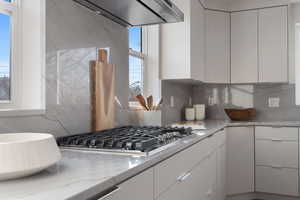 Kitchen featuring tasteful backsplash, light stone counters, white cabinetry, and range hood