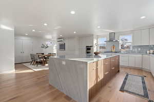 Kitchen with white cabinetry, a large island, wall chimney range hood, a wall mounted AC, and light white oak flooring