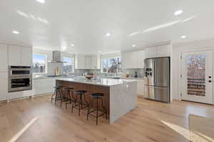 Kitchen featuring white cabinets, wall chimney range hood, light hardwood / wood-style floors, appliances with stainless steel finishes, and a kitchen island