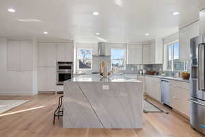 Kitchen featuring a center island, white cabinets, wall chimney exhaust hood, light hardwood / white oak floors, and stainless steel appliances