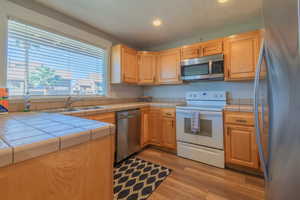 Kitchen featuring tile counters, sink, stainless steel appliances, light brown cabinetry, and light wood-type flooring