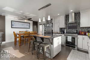 Kitchen featuring a center island, wall chimney exhaust hood, hanging light fixtures, stainless steel appliances, and white cabinets