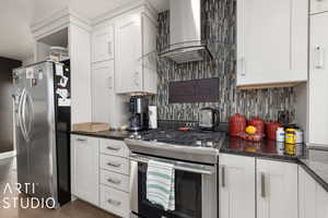 Kitchen with appliances with stainless steel finishes, tasteful backsplash, dark stone counters, wall chimney exhaust hood, and white cabinetry