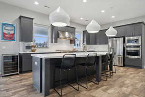 Kitchen featuring a kitchen island with sink, wine cooler, light wood-type flooring, appliances with stainless steel finishes, and decorative light fixtures