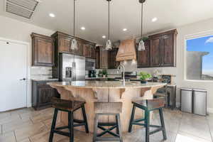 Kitchen featuring hanging light fixtures, light stone countertops, an island with sink, appliances with stainless steel finishes, and dark brown cabinets