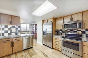 Kitchen featuring light stone counters, decorative backsplash, light brown cabinetry, appliances with stainless steel finishes, and light wood-type flooring