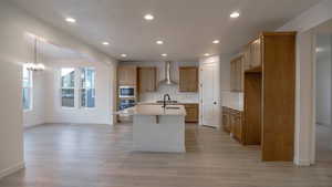 Kitchen with pendant lighting, light hardwood / wood-style floors, a center island with sink, built in microwave, and wall chimney exhaust hood