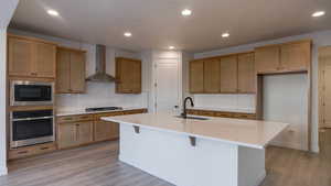 Kitchen with wall chimney range hood, sink, appliances with stainless steel finishes, light hardwood / wood-style floors, and an island with sink