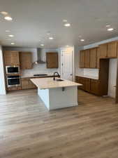 Kitchen featuring sink, wall chimney exhaust hood, stainless steel appliances, light hardwood / wood-style flooring, and a kitchen island with sink