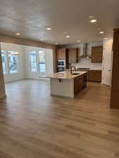 Kitchen featuring appliances with stainless steel finishes, wall chimney exhaust hood, a kitchen island with sink, pendant lighting, and light hardwood / wood-style floors