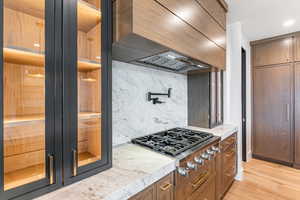 Kitchen featuring custom exhaust hood, a sauna / steam room, light stone counters, stainless steel gas cooktop, and light wood flooring