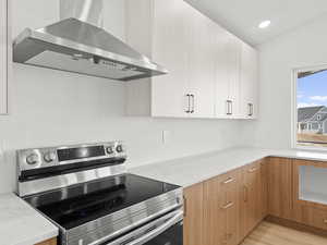 Kitchen with stainless steel electric range, white cabinets, wall chimney range hood, light stone countertops, and light hardwood / wood-style floors