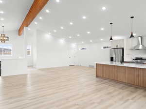 Kitchen featuring vaulted ceiling with beams, wall chimney exhaust hood, appliances with stainless steel finishes, decorative light fixtures, and light hardwood / wood-style floors