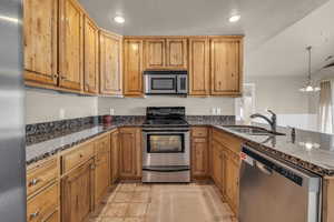 Kitchen featuring sink, dark stone counters, a textured ceiling, decorative light fixtures, and appliances with stainless steel finishes