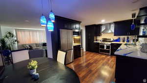 Kitchen with wall chimney exhaust hood, hanging light fixtures, tasteful backsplash, dark hardwood / wood-style flooring, and appliances with stainless steel finishes