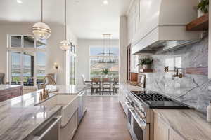 Kitchen with pendant lighting, backsplash, premium range hood, light stone counters, and stainless steel appliances