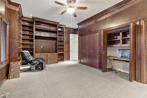 Unfurnished office / bedroom featuring wood walls, ceiling fan, ornamental molding, built in desk, and light colored carpet