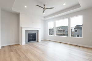 Unfurnished living room featuring ceiling fan, a raised ceiling, a tiled fireplace, and light hardwood / wood-style flooring