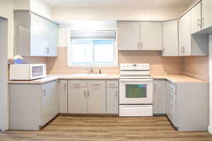 Kitchen features fresh paint, new light hardwood-style laminate flooring, light gray cabinets, new light fixtures, a new window with new blinds, white appliances, and sink