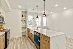 Kitchen with appliances with stainless steel finishes, pendant lighting, light stone countertops, light brown cabinets, and light hardwood / wood-style flooring