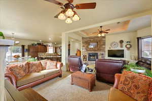 Carpeted living room with a fireplace, ceiling fan with notable chandelier, a raised ceiling, and plenty of natural light