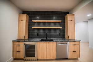 Bar featuring light brown cabinets, sink, beverage cooler, dark stone counters, and decorative backsplash