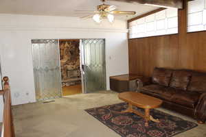 Living room with beam ceiling, wood walls, ceiling fan, and light colored carpet