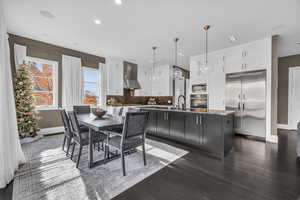 Interior space with wall chimney exhaust hood, built in appliances, decorative light fixtures, a kitchen island with sink, and white cabinets