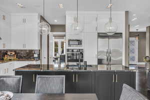 Kitchen with pendant lighting, built in appliances, dark stone countertops, tasteful backsplash, and white cabinetry