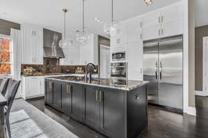 Kitchen with wall chimney exhaust hood, hanging light fixtures, built in appliances, an island with sink, and white cabinets