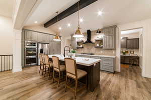Kitchen featuring wall chimney exhaust hood, hanging light fixtures, a center island with sink, gray cabinets, and stainless steel appliances
