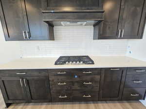 Kitchen featuring wall chimney exhaust hood, light wood-type flooring, tasteful backsplash, dark brown cabinets, and stainless steel gas cooktop