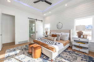 Bedroom with connected bathroom, ceiling fan, a barn door, a tray ceiling, and light wood-type flooring