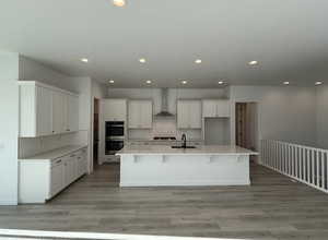 Kitchen with white cabinets, a spacious island, sink, wall chimney exhaust hood, and double oven