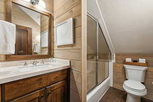 Full bathroom with wood walls, combined bath / shower with glass door, lofted ceiling, and toilet