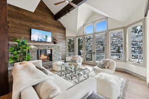 Living room with ceiling fan, a stone fireplace, high vaulted ceiling, wooden walls, and light wood-type flooring