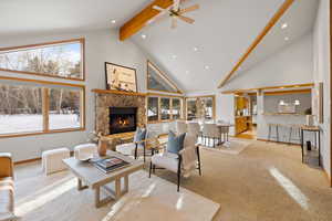 Carpeted living room featuring ceiling fan, beam ceiling, a stone fireplace, and high vaulted ceiling