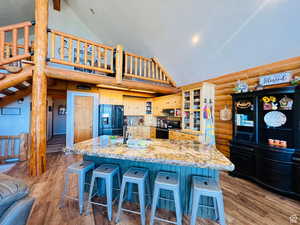 Kitchen featuring rustic walls, light hardwood / wood-style flooring, high vaulted ceiling, a center island with sink, and black appliances