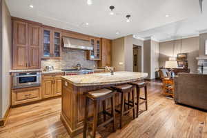 Kitchen featuring light stone countertops, light wood-type flooring, stainless steel appliances, wall chimney range hood, and an island with sink