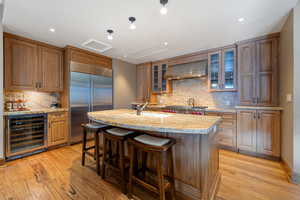 Kitchen featuring wine cooler, wall chimney exhaust hood, an island with sink, appliances with stainless steel finishes, and light hardwood / wood-style floors