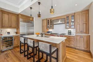 Kitchen featuring wall chimney range hood, an island with sink, appliances with stainless steel finishes, decorative light fixtures, and beverage cooler