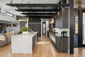 Kitchen featuring a high ceiling, wall chimney exhaust hood, light wood-type flooring, an island with sink, and light stone counters