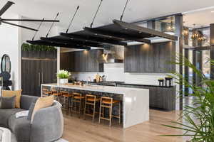 Kitchen with a kitchen island with sink, wall chimney range hood, light stone countertops, light wood-type flooring, and dark brown cabinets