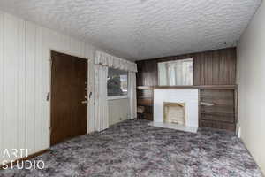 Unfurnished living room with dark colored carpet, a textured ceiling, wooden walls, and a brick fireplace