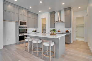 Kitchen featuring gray cabinets, wall chimney exhaust hood, appliances with stainless steel finishes, a kitchen bar, and recessed lighting