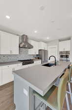 Kitchen with a kitchen bar, appliances with stainless steel finishes, a kitchen island with sink, wall chimney range hood, and white cabinetry