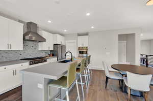 Kitchen with wall chimney exhaust hood, decorative backsplash, an island with sink, white cabinetry, and stainless steel appliances