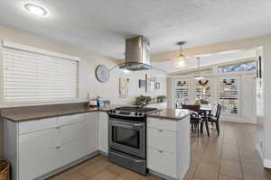 Kitchen featuring pendant lighting, kitchen peninsula, stainless steel gas stove, island range hood, and white cabinetry