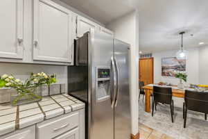 Kitchen with stainless steel fridge, light tile patterned floors, tile countertops, and white cabinetry