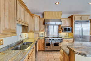 Kitchen featuring sink, wall chimney exhaust hood, light brown cabinets, premium appliances, and light stone counters